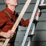 cropped view of repairman in orange uniform climbing with wooden board in hand on ladder in garage